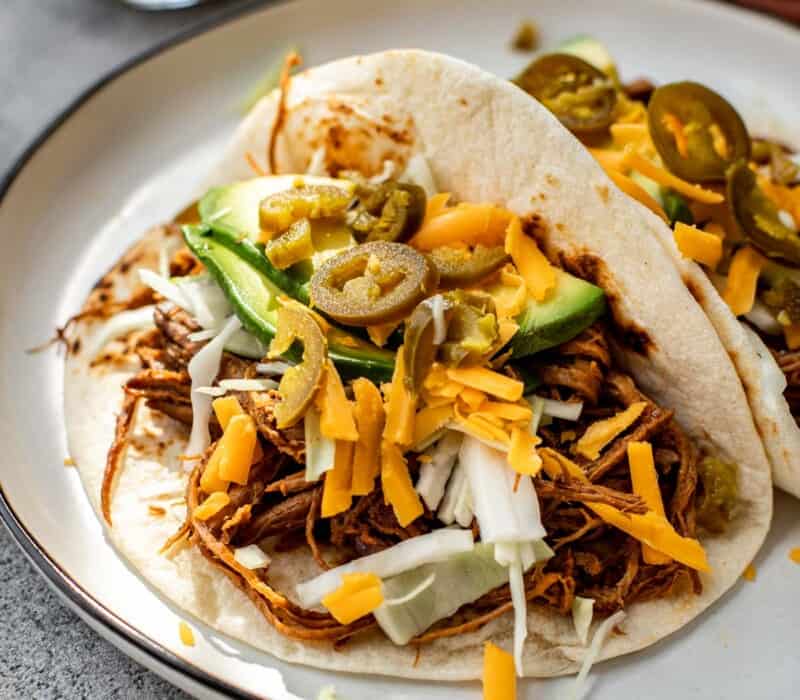Shredded beef tacos on a plate topped with shredded cheese, avocados, pickled jalapenos, and shredded cabbage.