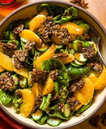 little gem lettuce leaves topped with orange segments, chunks of granola and sliced cucumbers in a large buttery-yellow serving bowl.