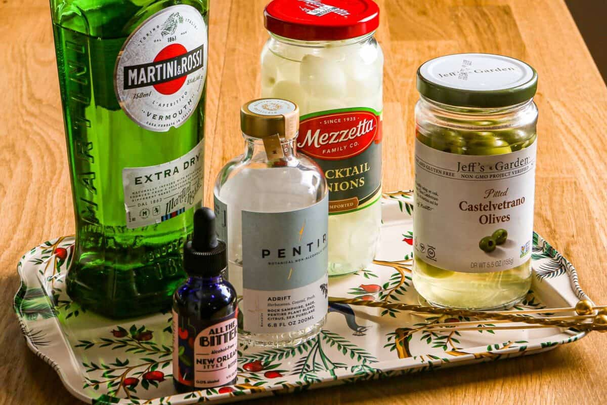 ingredients for a non alcoholic martini set on a floral tray. Including non alcoholic gin, vermouth, non alcoholic bitters, olives, and cocktail onions.
