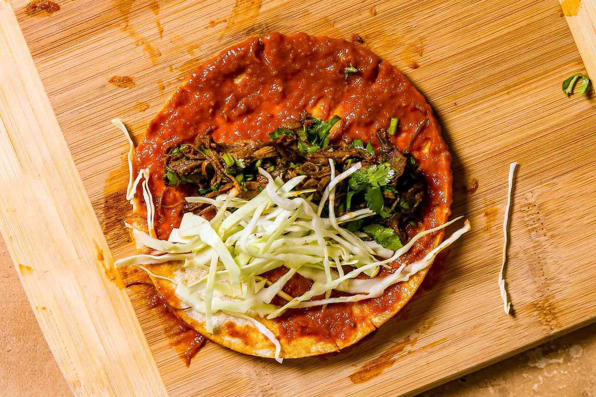 A small corn tortilla coated in a red enchilada sauce with shredded beef and shredded cabbage arranged down the middle.