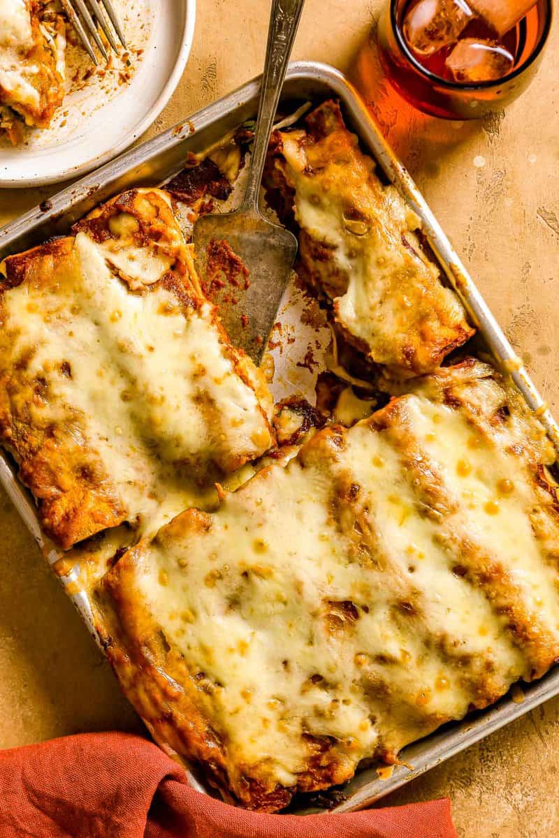 A small sheetpan of cheese-covered enchiladas with a few taken out and a serving utensil set in the pan.