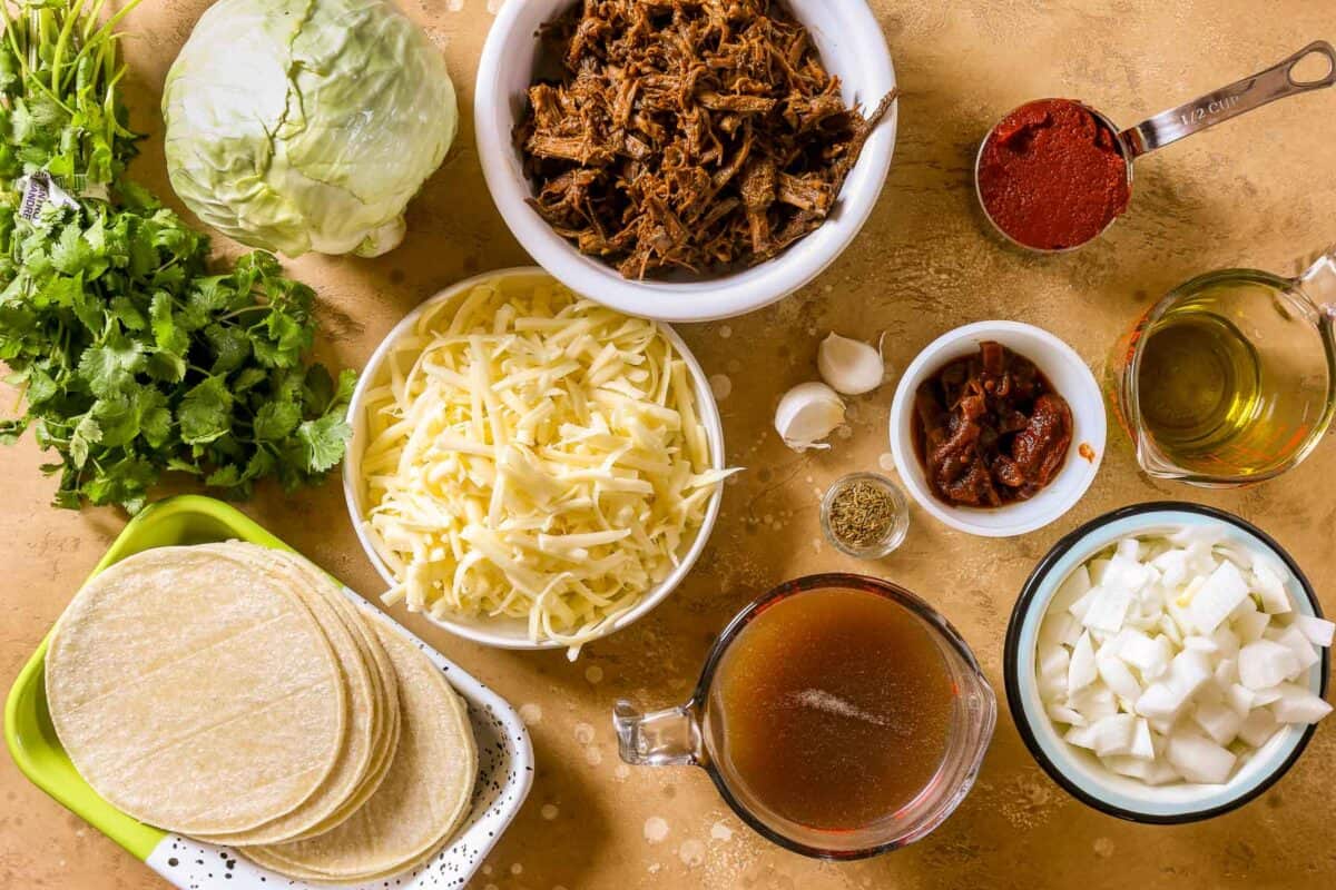 The ingredients for shredded beef enchiladas set out—garlic, olive oil, tomato paste, chipotle peppers, broth, onions, shredded beef, shredded cheese, head of cabbage, cilantro and tortillas.