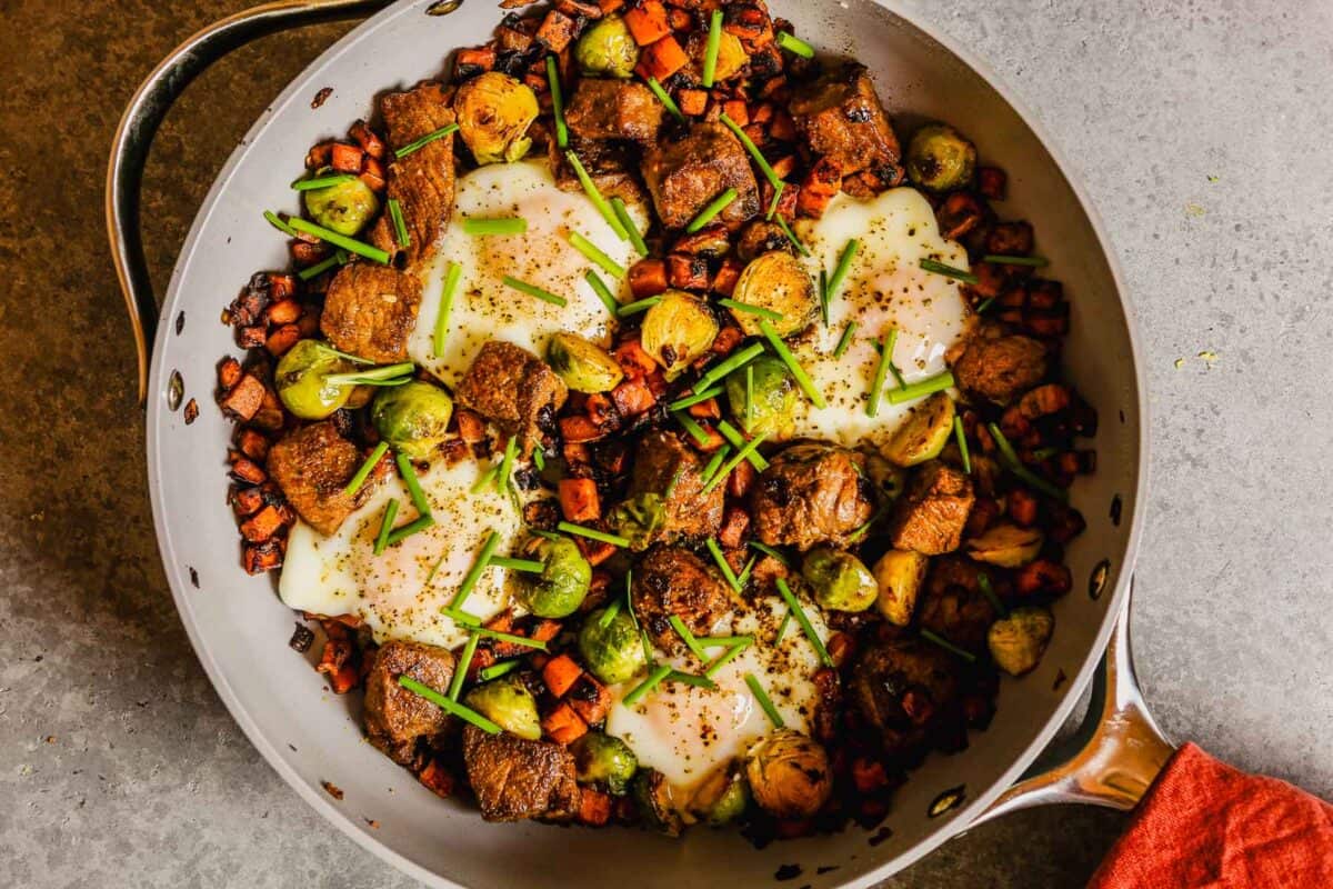 Cubes of cooked steak, diced cooked sweet potato, onion and cooked brussels sprouts in a nonstick skillet with 4 cooked eggs nestled into the breakfast hash.