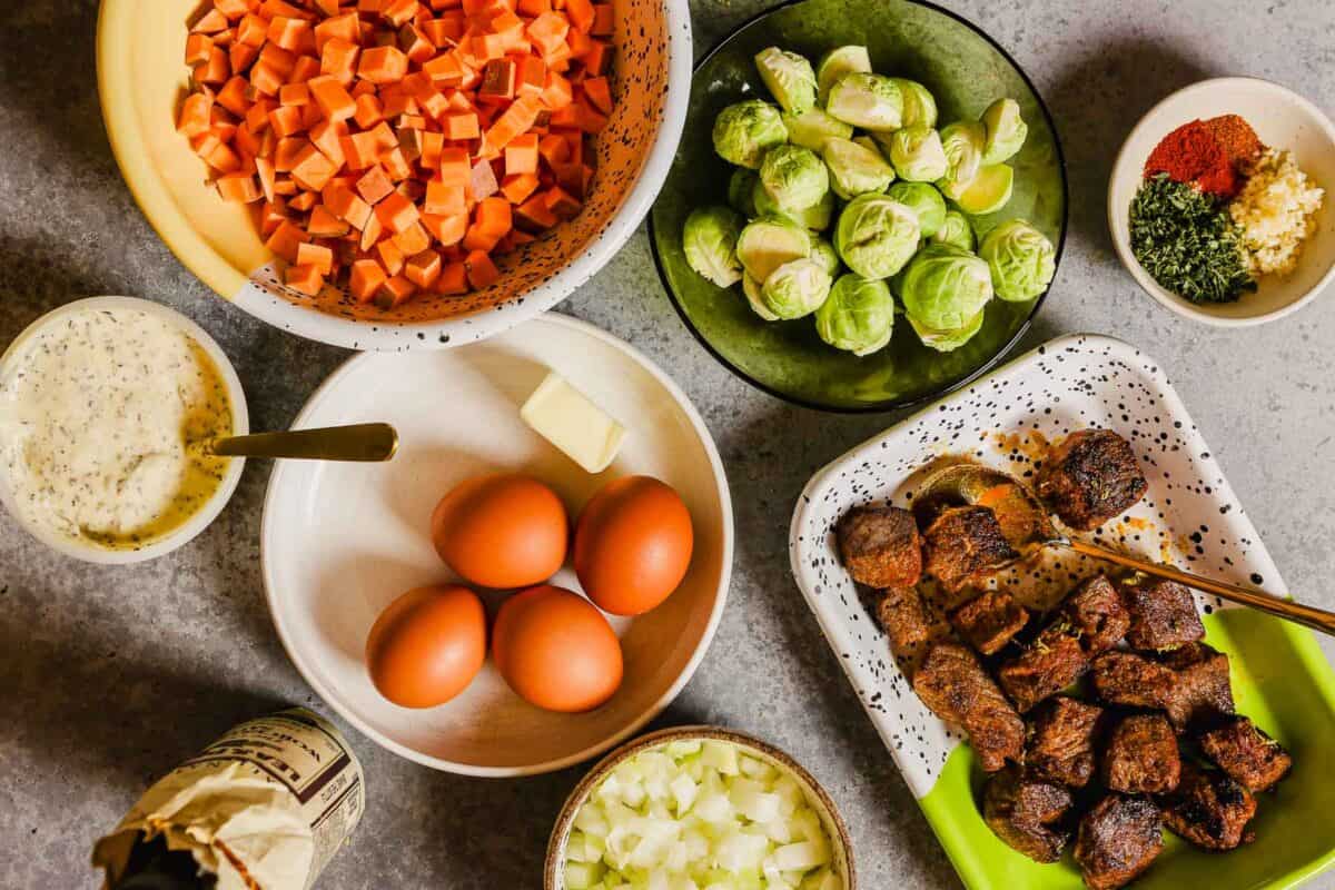 diced sweet potatoes, halved brussels sprouts, spices, eggs, cooked steak, onions, sauce and worcestershire set out on a counter. 