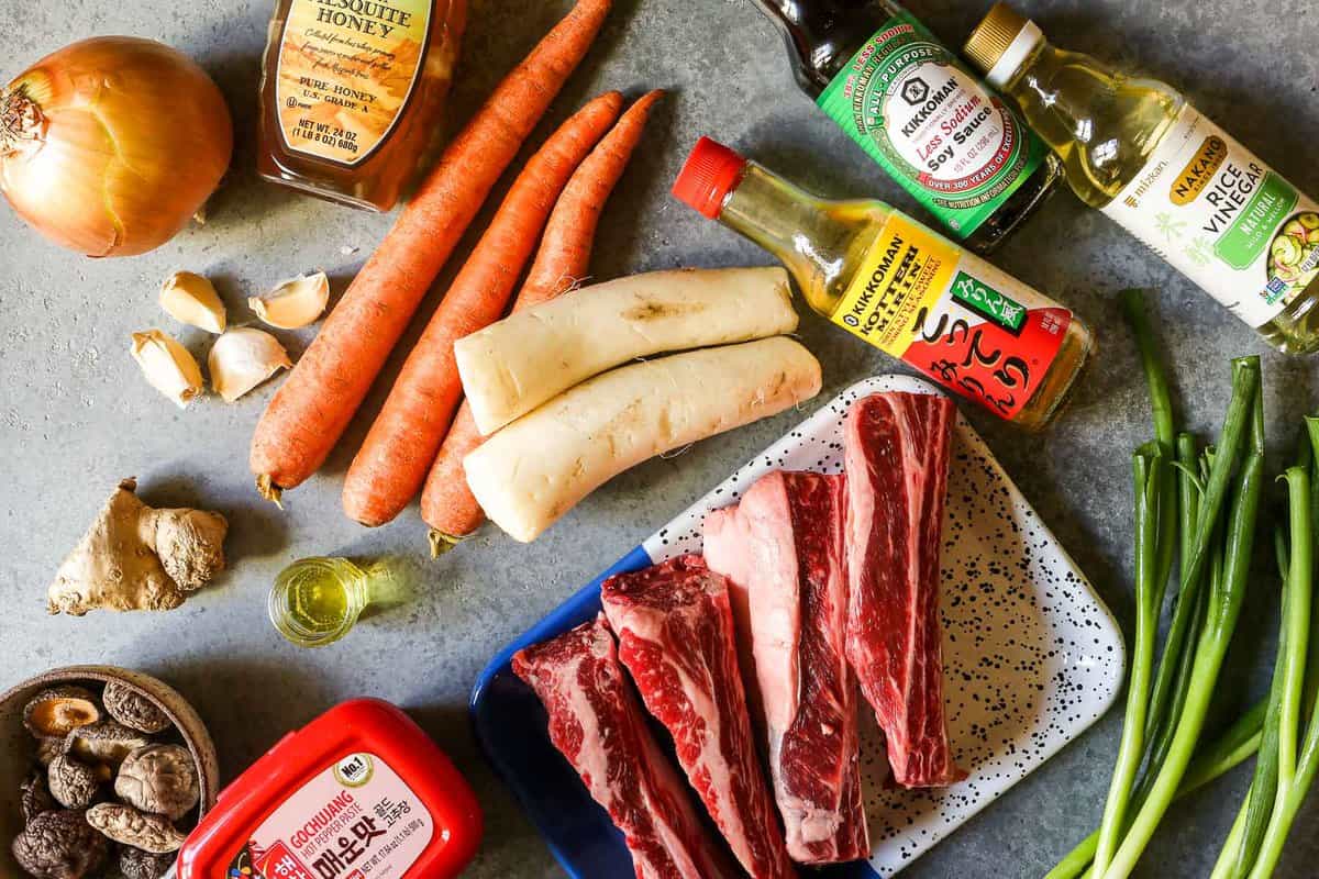 Beef shortribs, gochujang, soy sauce, mirin, carrots, daikon, oil. garlic, honey, rice vinegar, and scallions set out on a counter.
