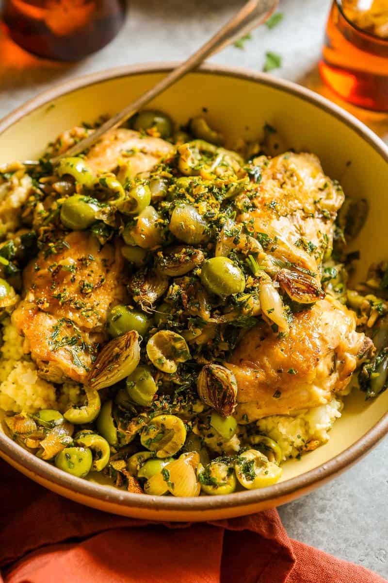 Chicken martini thighs in a large serving bowl with green olives, browned pearl onions, lemon, parsley and couscous.