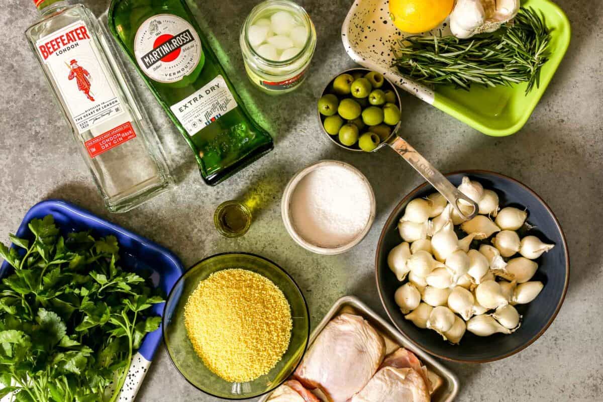 A bottle of gin, a bottle of vermouth, a lemon, bunch of parsley, pearl onions, skin-on chicken thighs, salt, oil, olives, rosemary, and garlic set out on a counter.