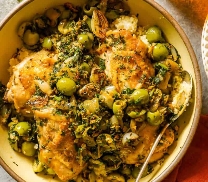 Chicken martini thighs in a large serving bowl with green olives, browned pearl onions, lemon, parsley and couscous.