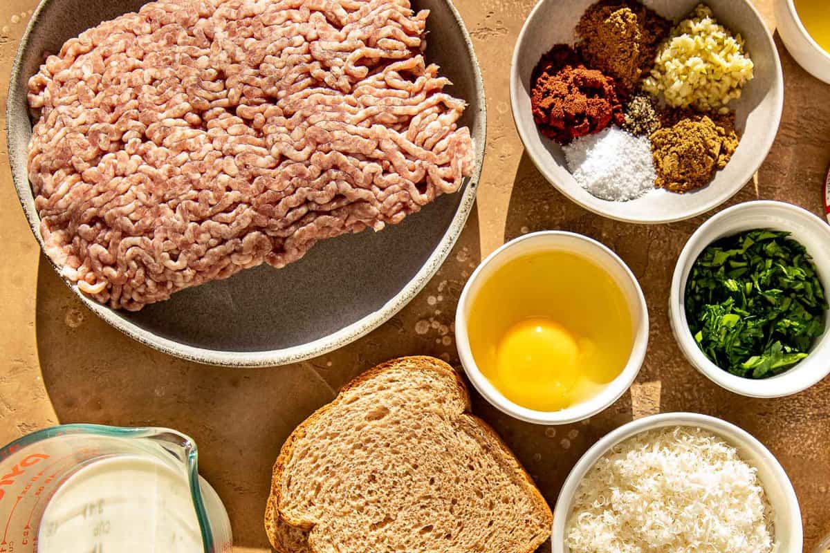 Ground pork, spices, garlic, egg, milk, bread, cheese and olive oil set out on a counter. 