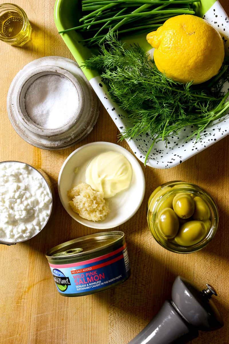 A can of salmon, drained green olives, pepper grinder, mayonnaise, horseradish, cottage cheese, salt, olive oil, cill and lemon set out on a counter.