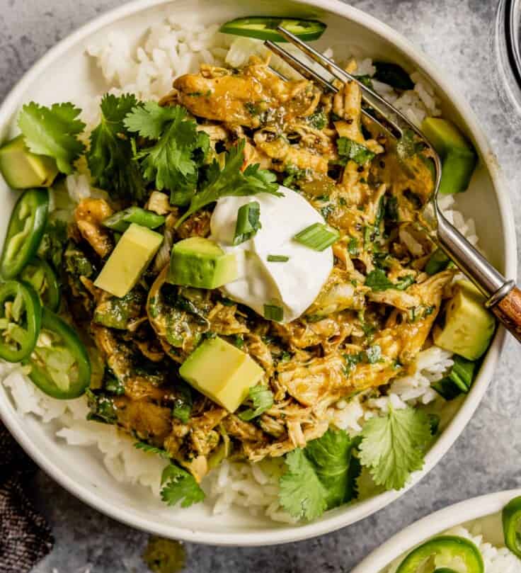 Shredded salsa verde chicken served over rice in a shallow white bowl topped with avocado, cilantro and jalapenos.