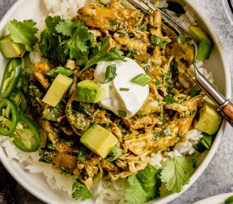 Shredded salsa verde chicken served over rice in a shallow white bowl topped with avocado, cilantro and jalapenos.