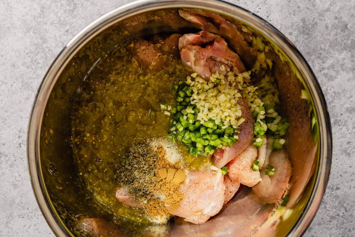 Raw chicken breast and chicken thighs in a pot covered in salsa verde, garlic and minced jalapeno.