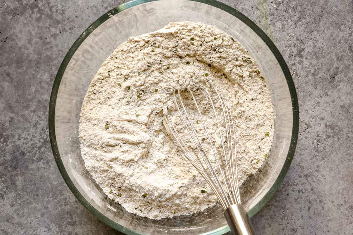 All purpose flour, rye flour, dried chives, and dried dill in a glass mixing bowl with a whisk set in it.