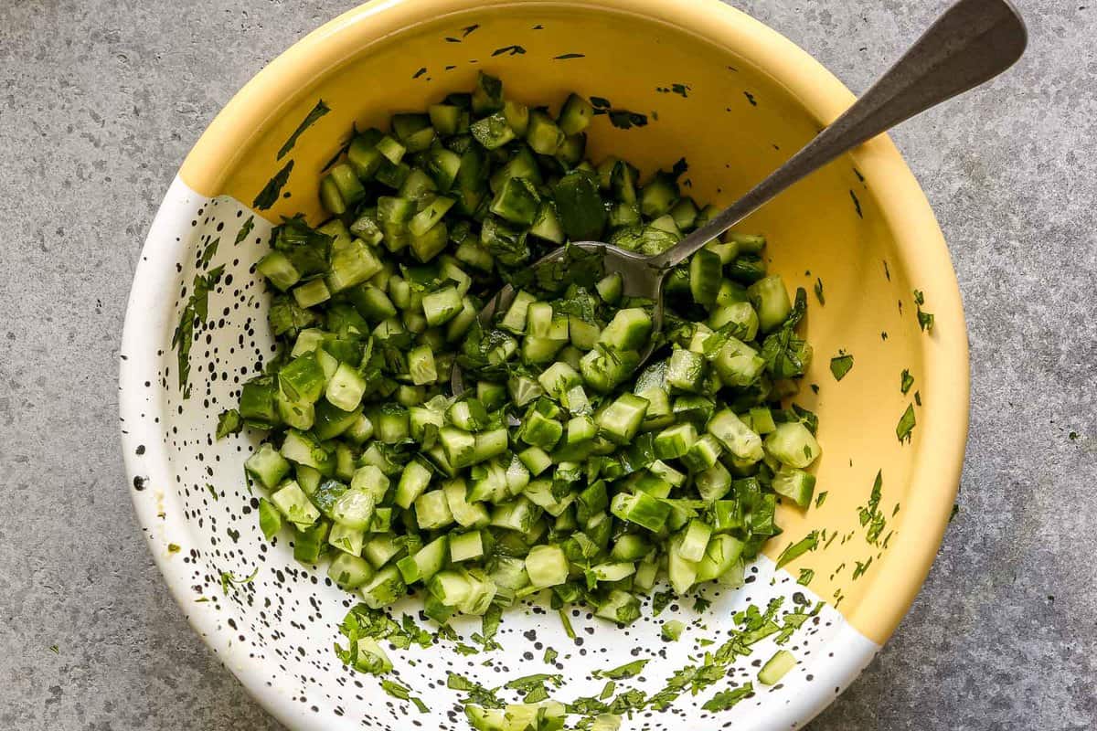 Diced cucumber tossed in minced cilantro in a metal mixing bowl.