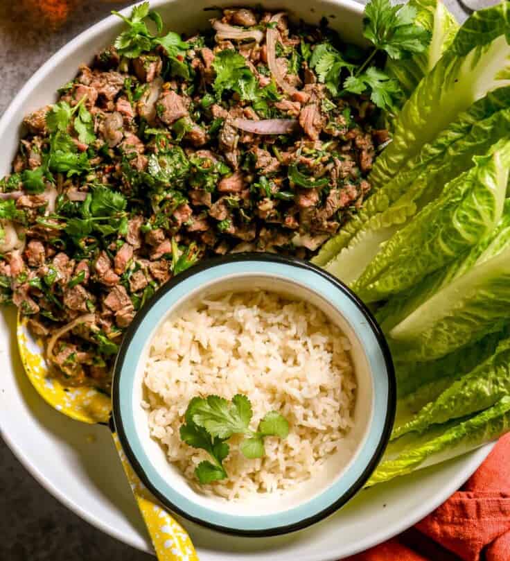 Large white serving bowl filled with meat salad, a small bowl of rice and lettuce leaves.