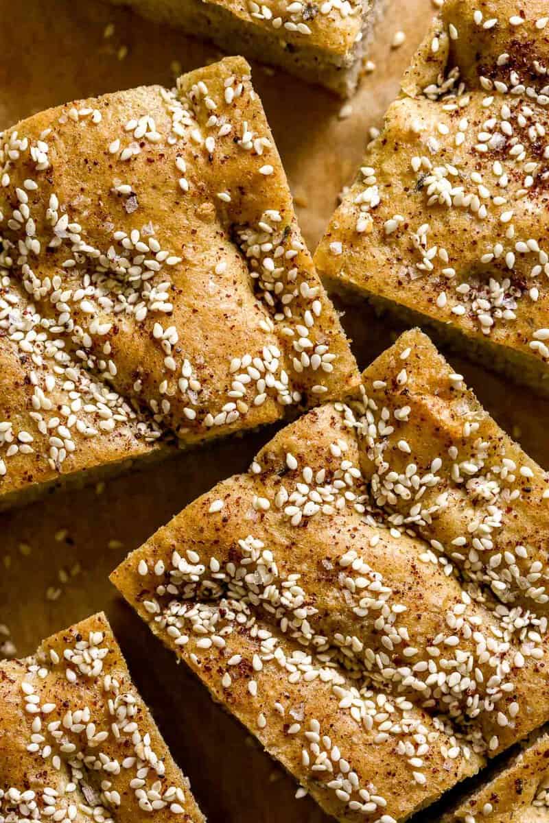 Large pieces of no knead focaccia bread on parchment paper. Bread topped with sesame seeds, salt and sumac.