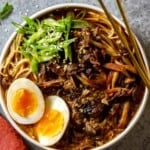 Beef ramen in a shallow white bowl topped with soft boiled egg halves, sliced scallions, browned mushrooms and sesame seeds. Chop sticks set on the bowl.