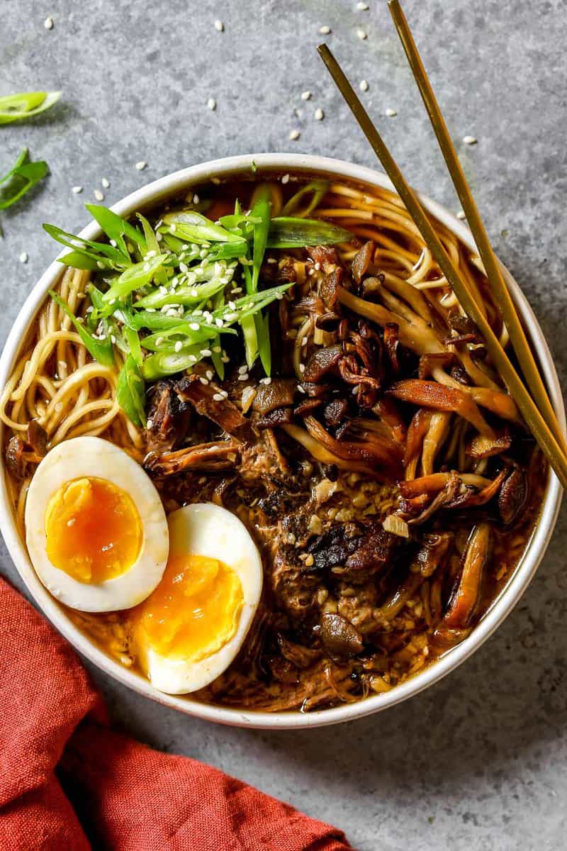 Beef ramen in a shallow white bowl topped with soft boiled egg halves, sliced scallions, browned mushrooms and sesame seeds. Chop sticks set on the bowl.