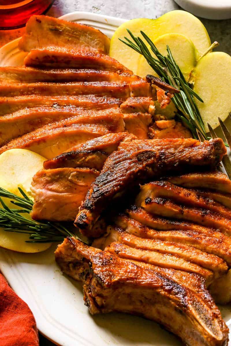 Sliced bone-in brined pork chop on a large white platter with apples and rosemary set around it.