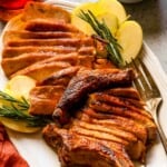 Sliced bone-in pork chop on a large white platter with apples and rosemary set around it.