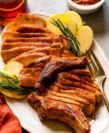 Sliced bone-in pork chop on a large white platter with apples and rosemary set around it.
