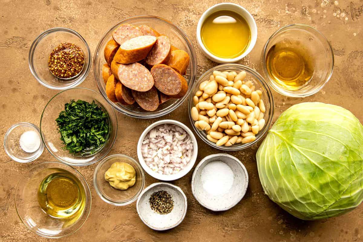 Mustard, herbs, sausage, cabbage, oil, honey, shallots and beans set out on a counter in preparation for making Cabbage and Sausage recipe.