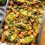 Cabbage, sausage and white beans on a baking sheet with herby sauce spooned over top.