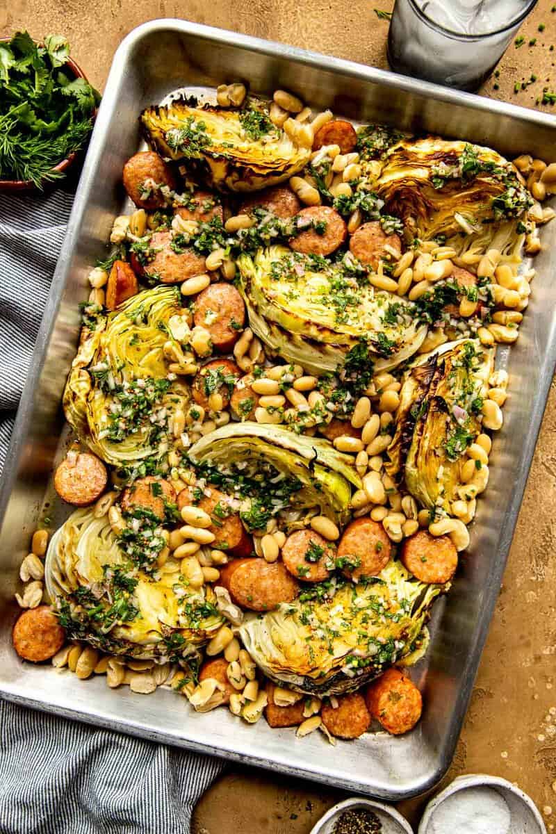 Cabbage, sausage and white beans on a baking sheet with herby sauce spooned over top.