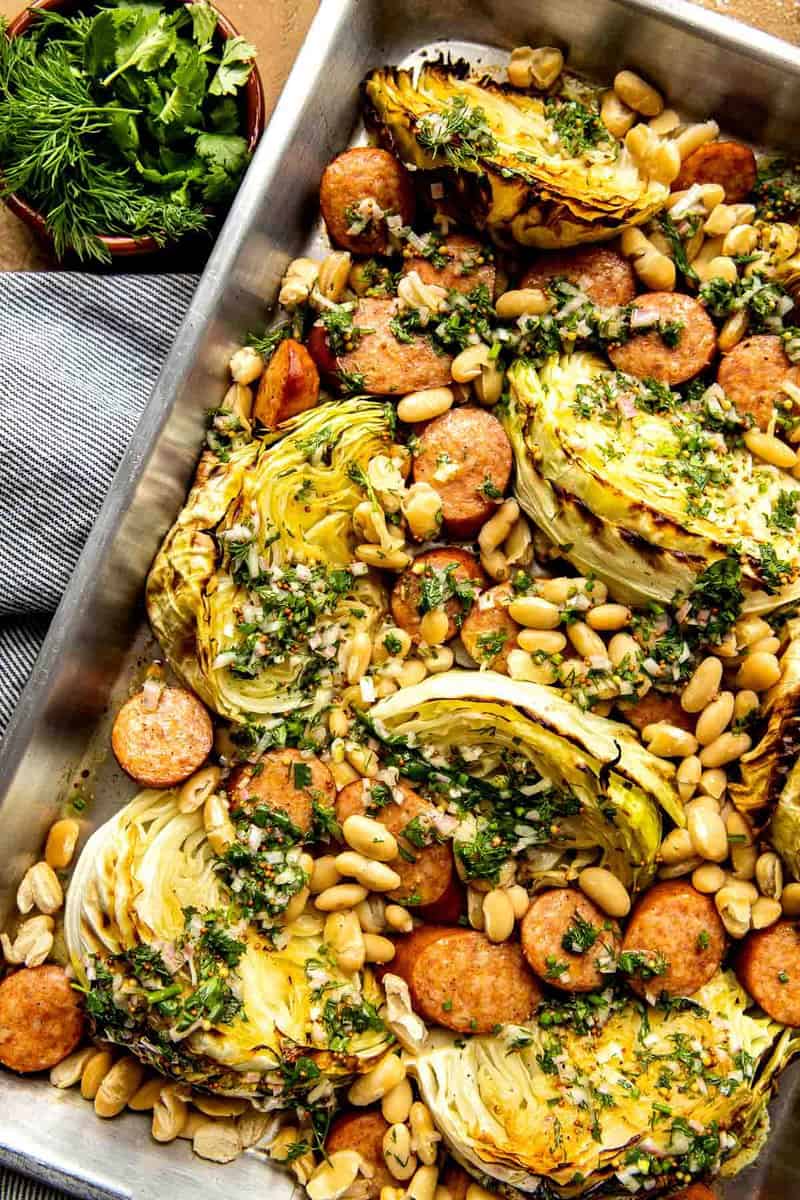 Cabbage, sausage and white beans on a baking sheet with herby sauce spooned over top.