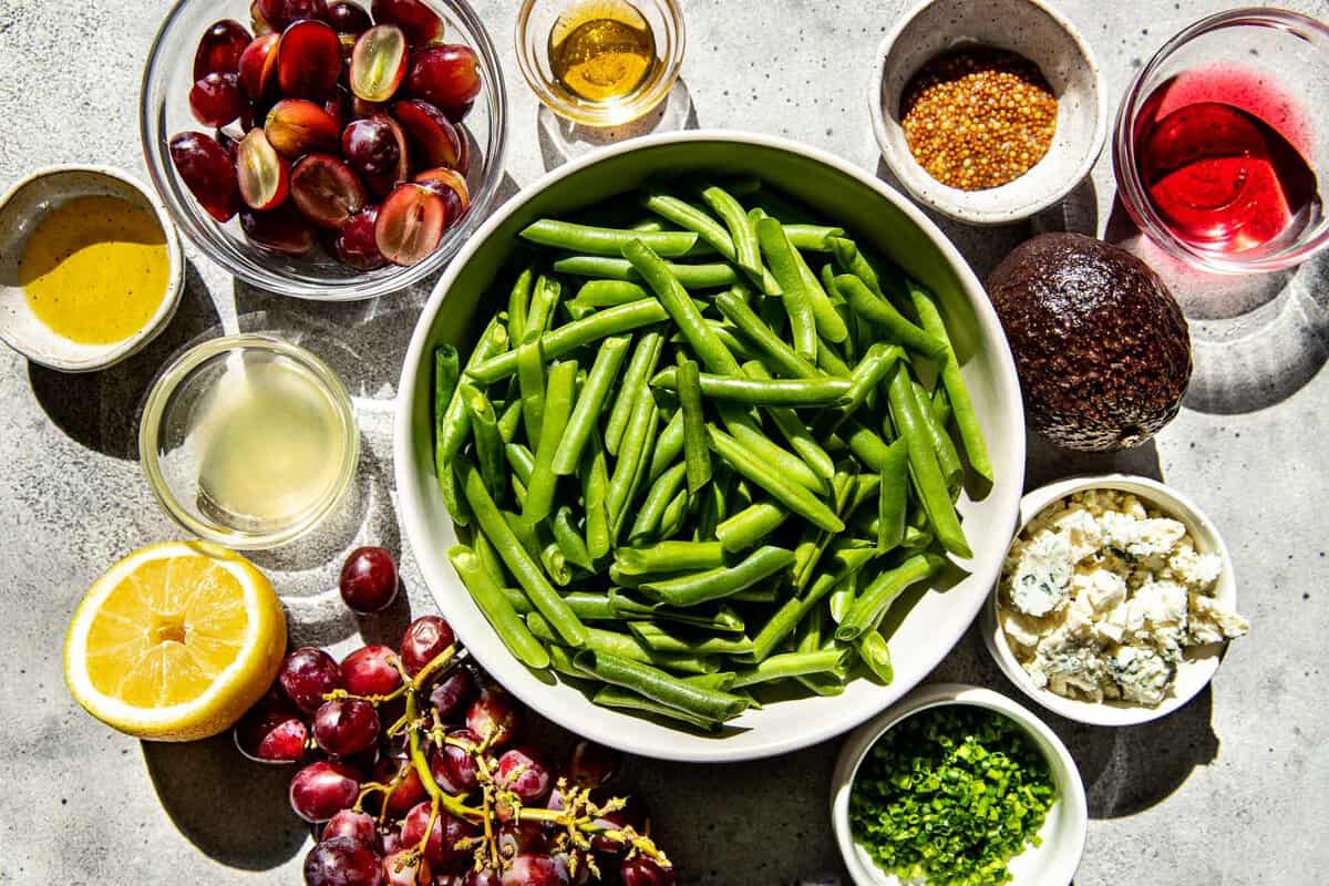 blanched green beans, grapes, avocado, chives, blue cheese, oil, mustard, vinegar, and lemon set out on the counter. 