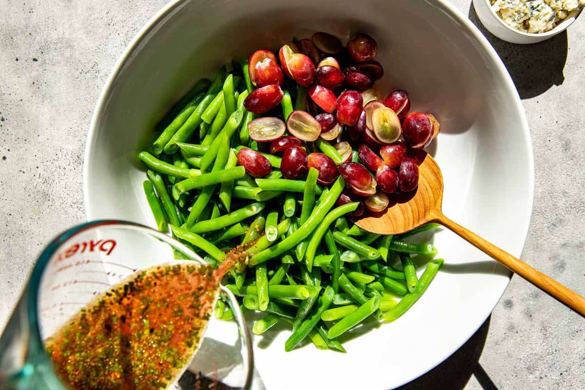 vinaigrette getting poured into a bowl with green beans and grapes.