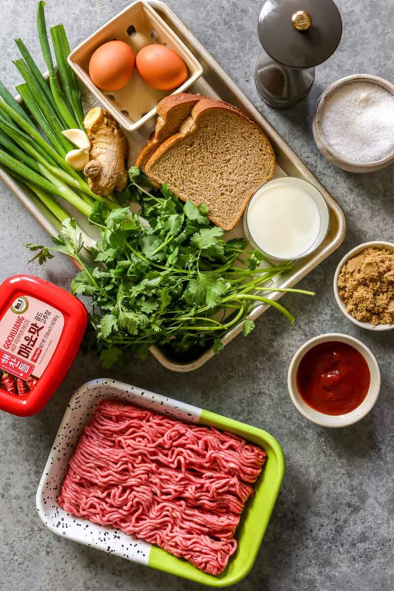 Ground beef, gochujang, scallions, cilantro, ginger, garlic, bread, milk, ketchup, brown sugar and eggs set out on a counter in preparation for making mini meatloaf.
