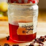 Hibiscus syrup in a large jar set on a wood-top counter with dried hibiscus leaves set around it.