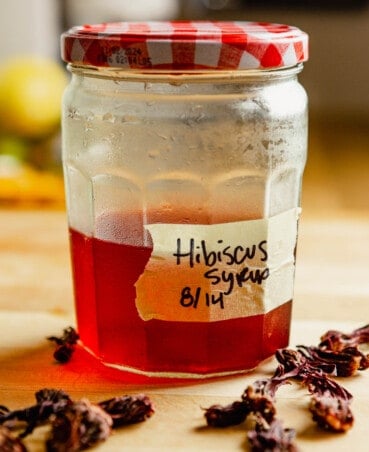 Hibiscus syrup in a large jar set on a wood-top counter with dried hibiscus leaves set around it.
