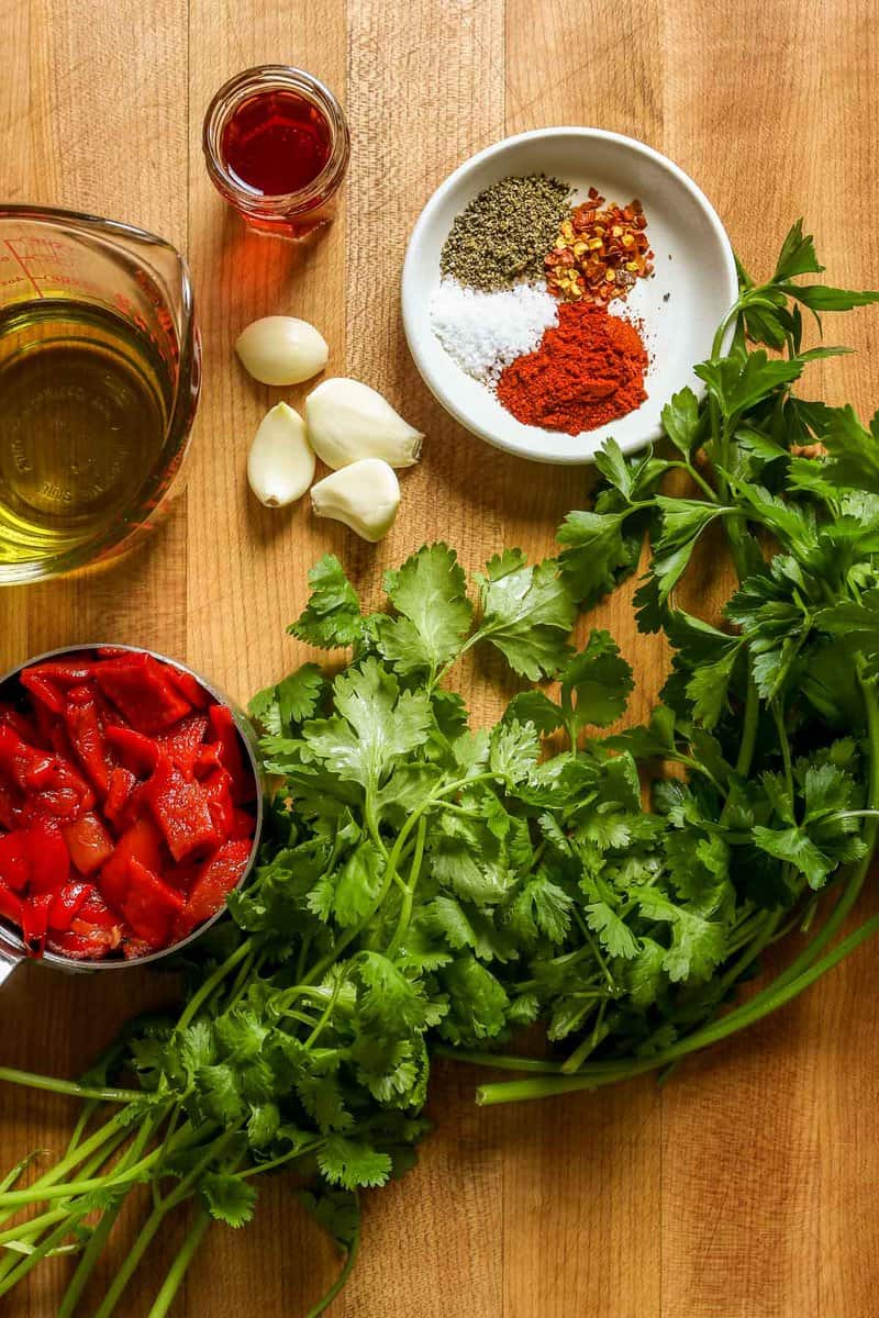 Olive oil, garlic cloves, roasted red peppers, cilantro, parsley, spices and vinegar set out on a counter.