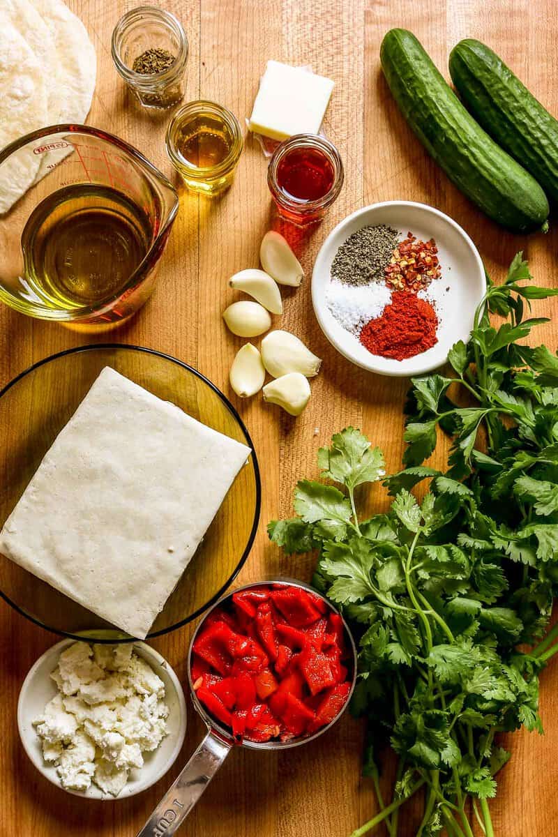 Tofu, tortillas, cheese, olive oil, garlic cloves, roasted red peppers, cilantro, parsley, spices and vinegar set out on a counter.