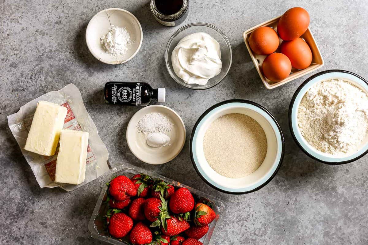 Butter, fresh strawberries, sour cream, sugar, vanilla, eggs, flour, baking powder and salt set out on a counter. 