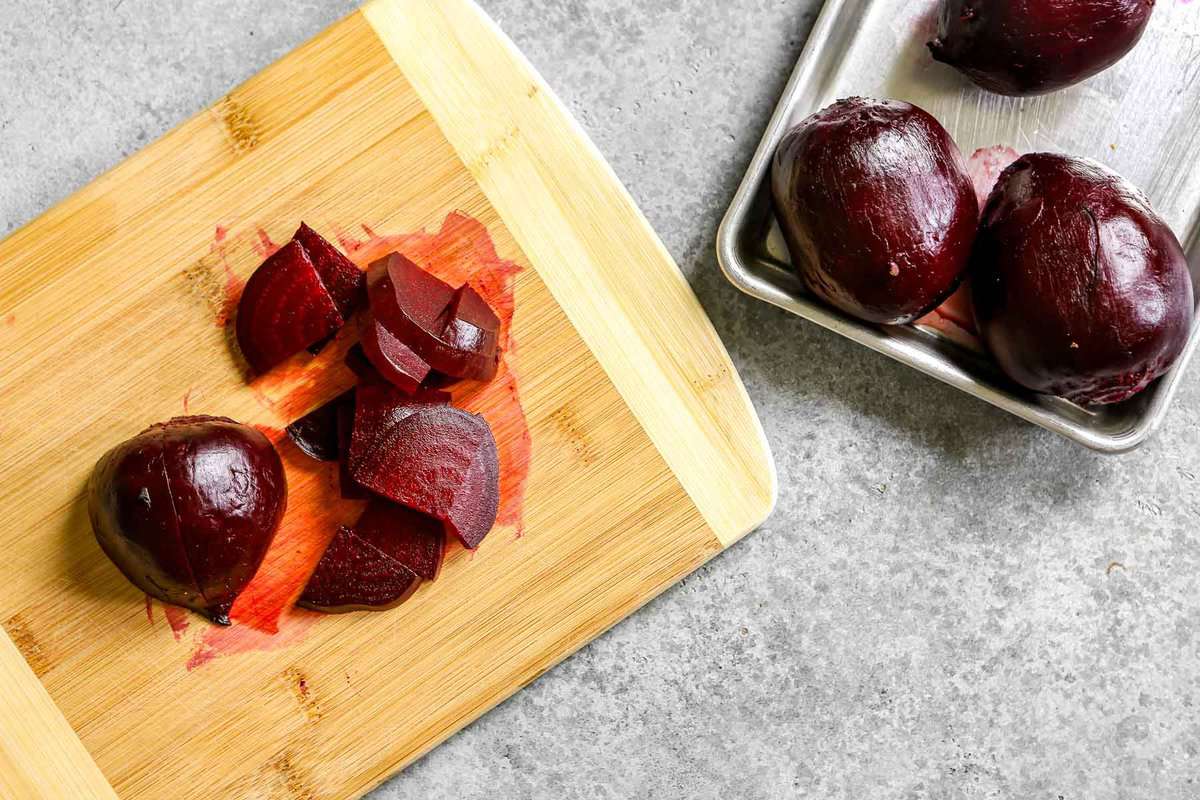 Roasted beets on a sheet pan with one beet on a wood cutting board getting sliced.