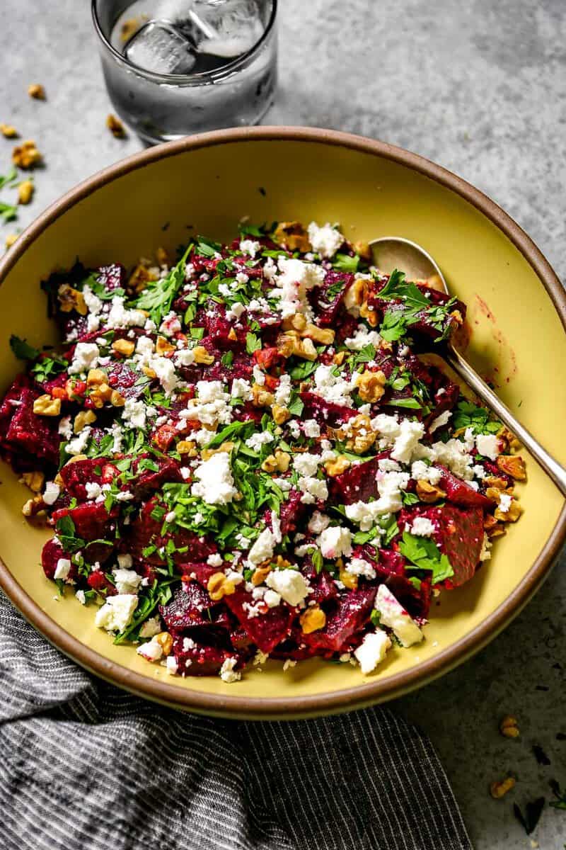 Beetroot salad with crumbled feta, parsley, and walnuts in a yellow ceramic bowl with two silver serving spoons.