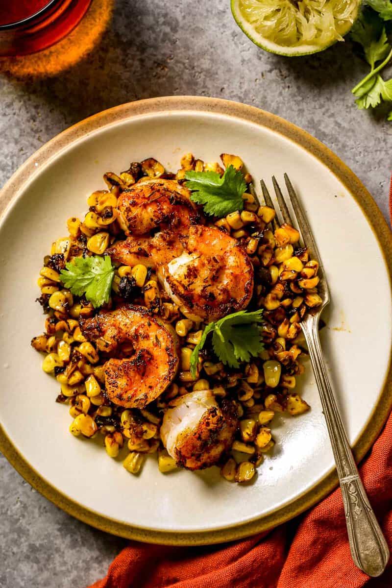 Blackened shrimp set over a bed of blackened sweet corn on a white dinner plate with a silver fork set on plate. Orange water glass, juiced lime half and fresh cilantro set around the plate. 