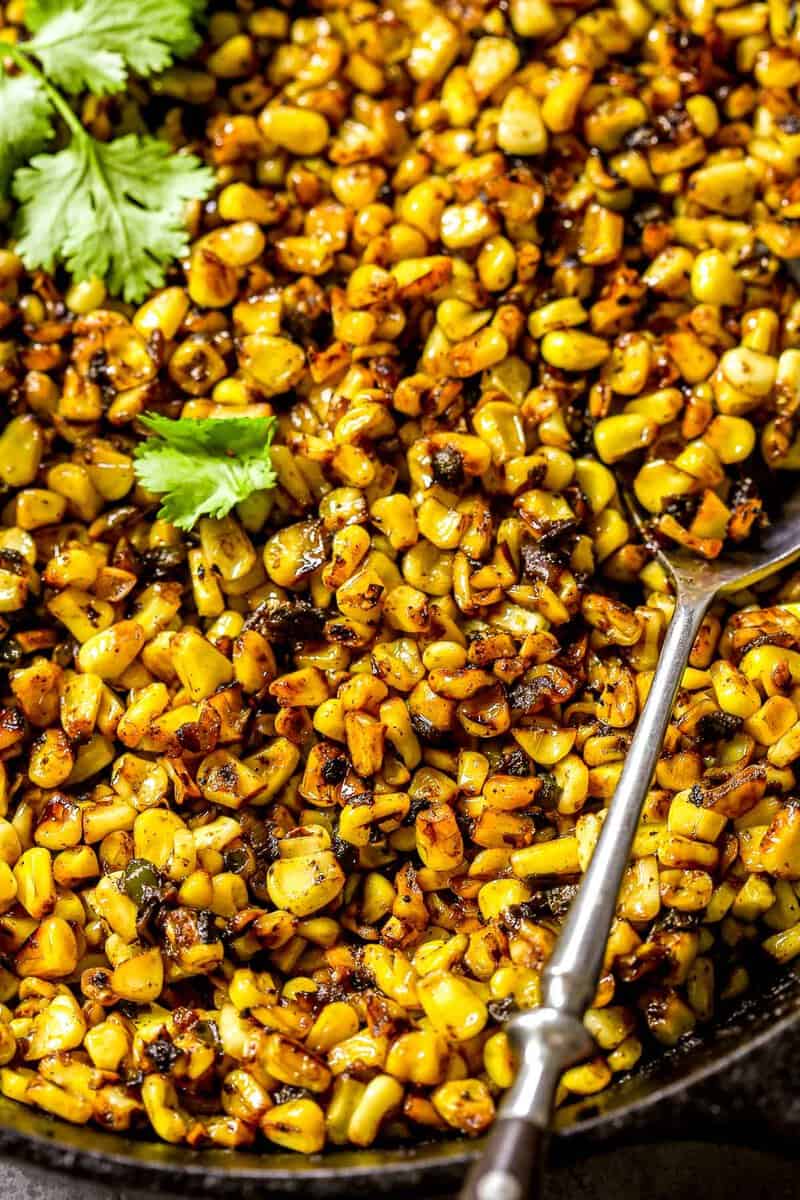 Blackened sweet corn in a cast-iron skillet with fresh cilantro scattered over top and a silver spoon set in it.
