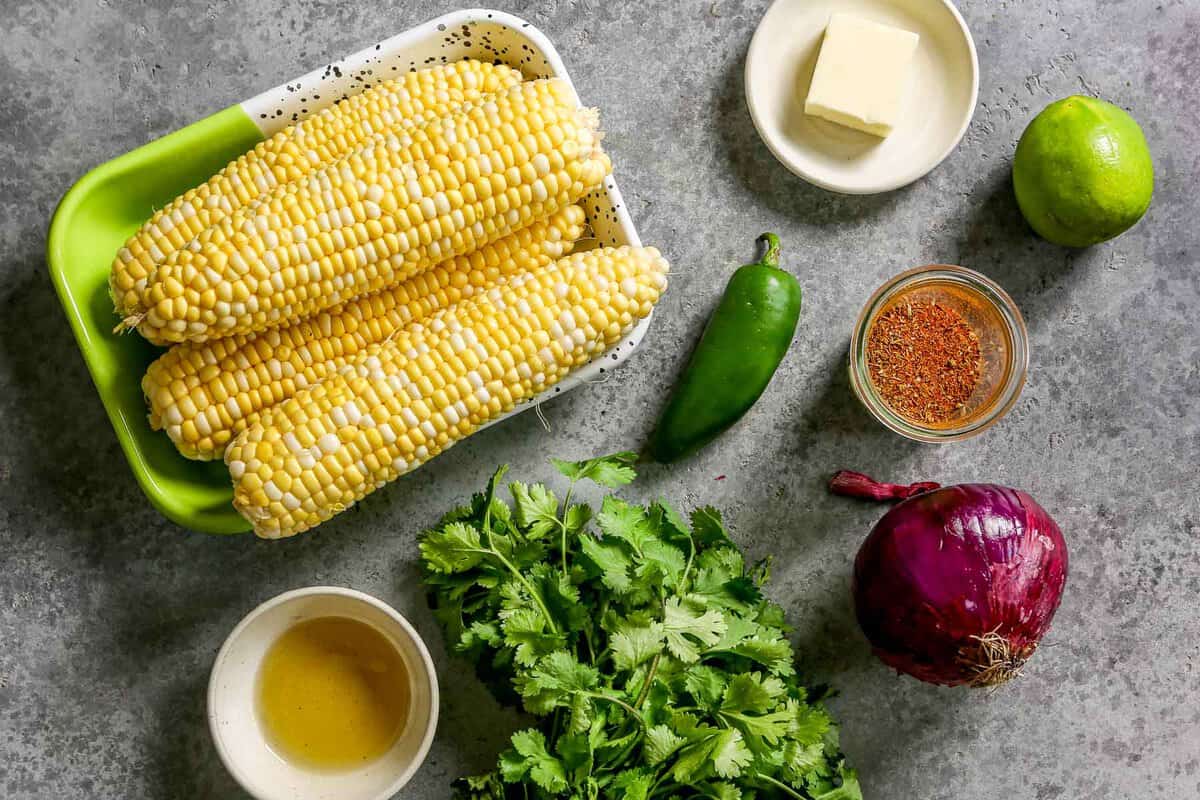 Four ears of shucked sweet corn on a speckled metal platter, a bowl of oil, fresh cilantro, a jalapeno, red onion, Cajun seasoning, butter, and lime set out on a counter. 