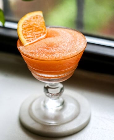 Frozen Aperol Spritz in a coupe glass with an orange slice set in cocktail. Set on a white counter with greenery in the background.