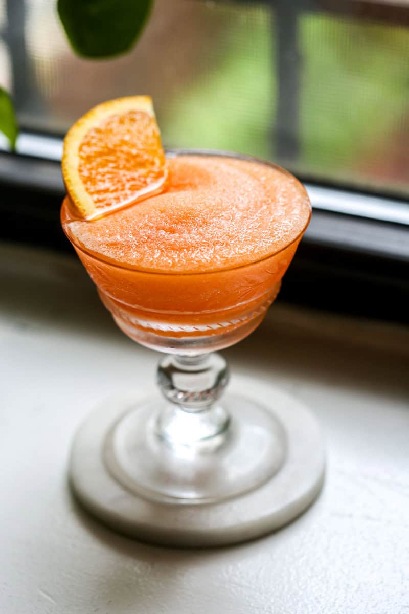 Frozen Aperol Spritz in a coupe glass with an orange slice set in cocktail. Set on a white counter with greenery in the background.