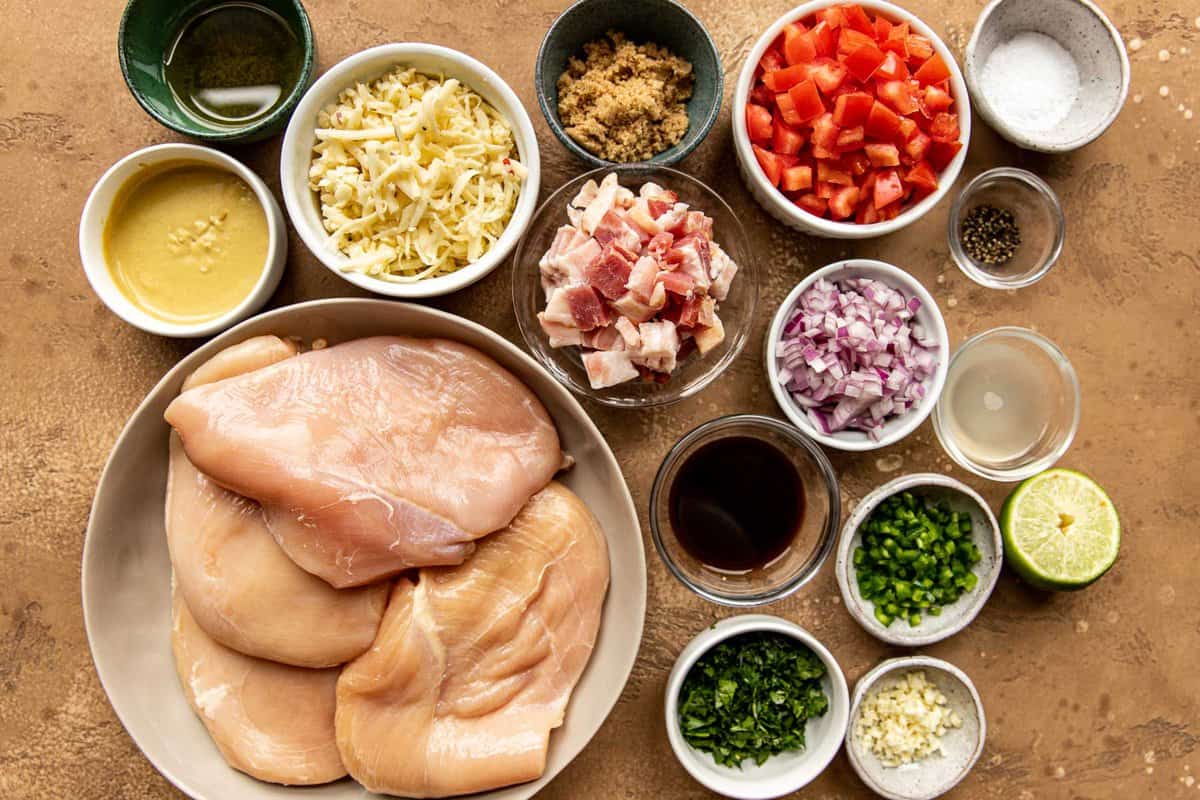 Raw chicken breasts, shredded cheese, Dijon, brown sugar, diced uncooked bacon, chopped cilantro, chopped jalapeno, chopped onion, chopped red onion, minced garlic, Worcesterhsire, salt and pepper all measured out and in bowls set on a counter.