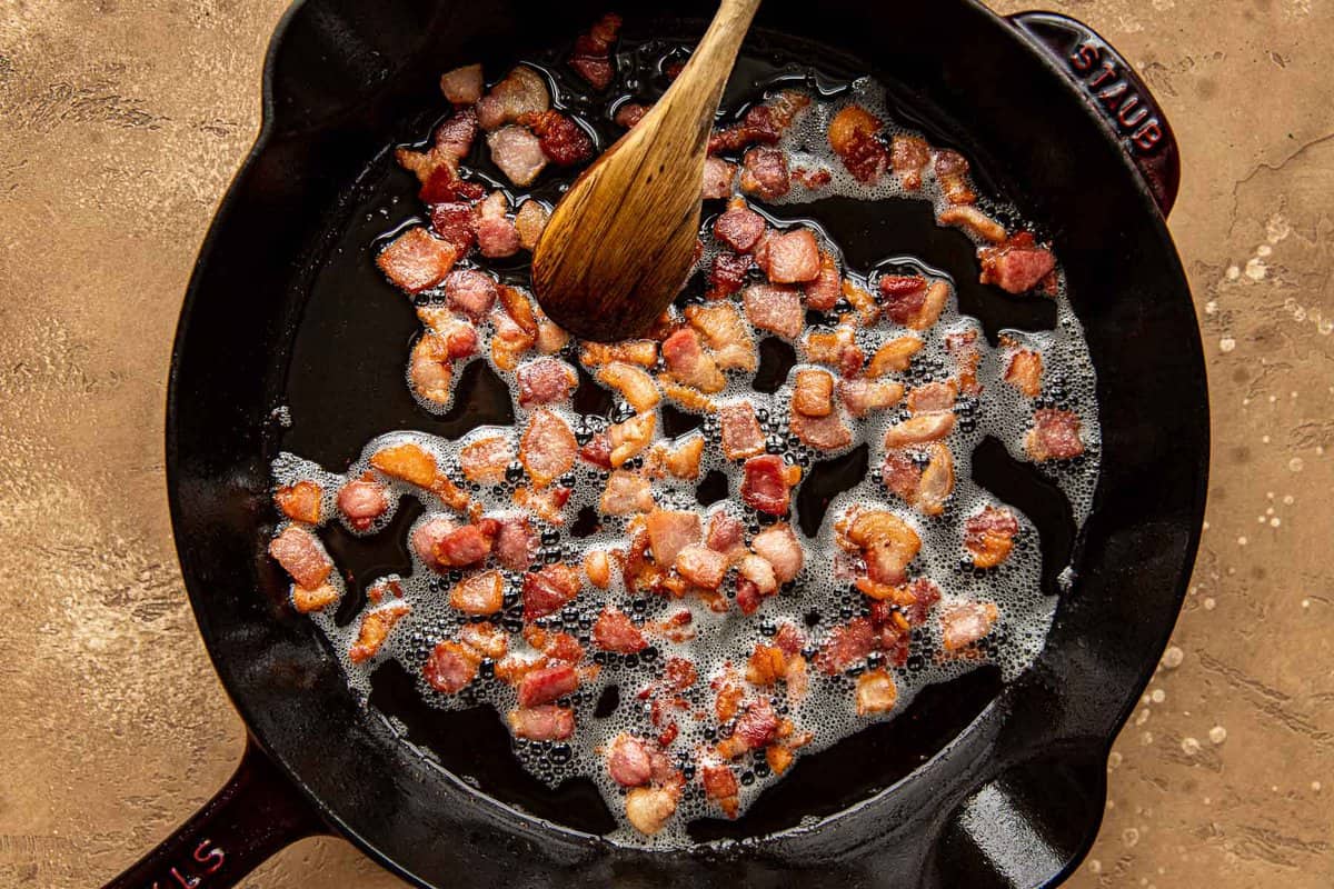 Diced bacon cooking in a cast-iron skillet with a wooden spoon stirring the bacon.