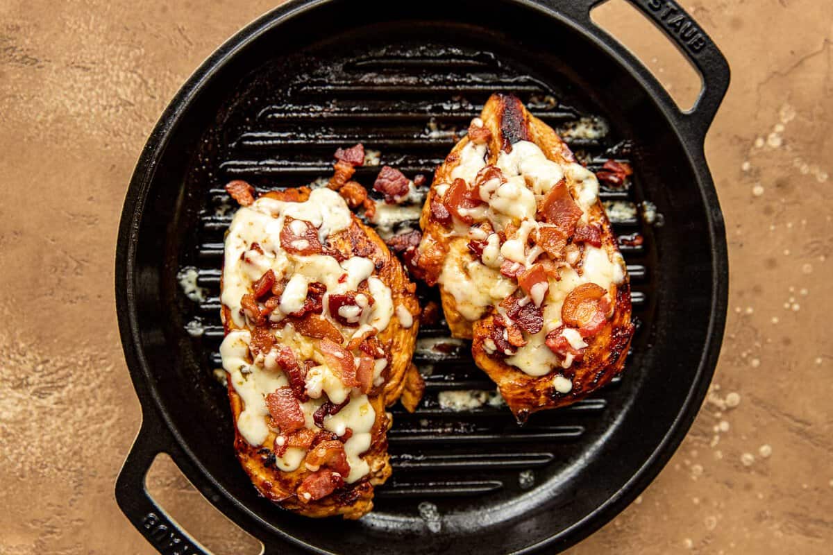 Two chicken breasts topped with melty cheese and crumbled bacon, cooking on a grill pan.