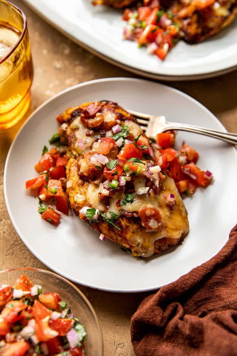 Grilled chicken breast topped with melted cheese, crispy bacon bits and pico de gallo on a white dinner plate with a fork set on plate and a yellow glass set to the top right.