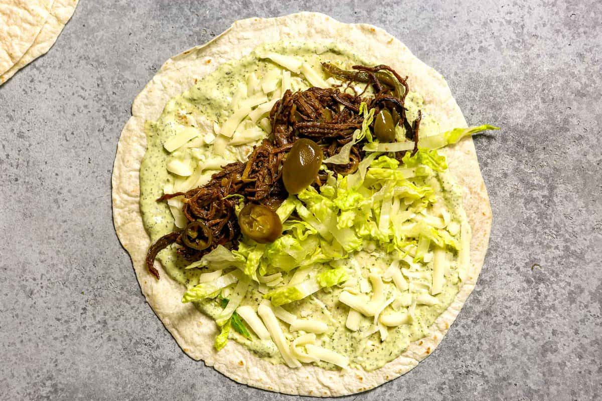 Flour tortilla set on a work surface with burrito filling on it—chimichurri mayo spread on the tortilla, shredded cheese sprinkled over top, shredded beef arranged in a line, shredded lettuce arranged next to beef and pickled jalapeno slices on top of the filling.